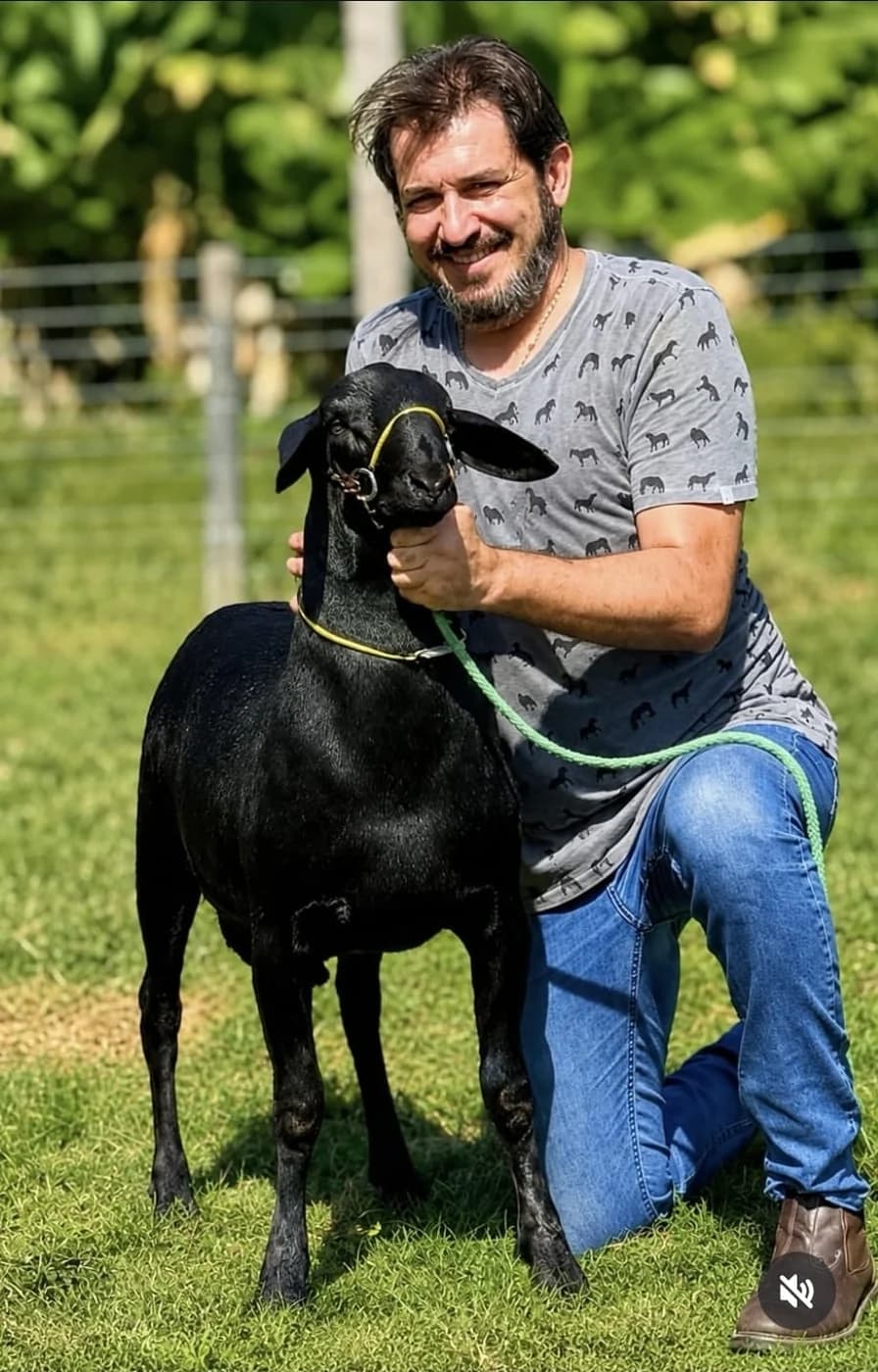 Augusto Cesinha ajoelhado no pasto, segurando pelo cabresto uma ovelha Santa Inês preta