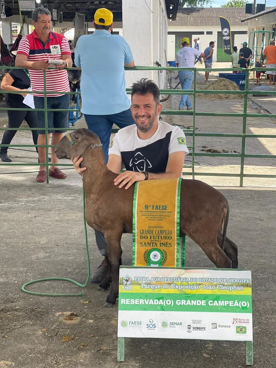 Augusto Cesinha abraçado a uma ovelha Santa Inês marrom com faixa de premiação, ao lado da placa da feira