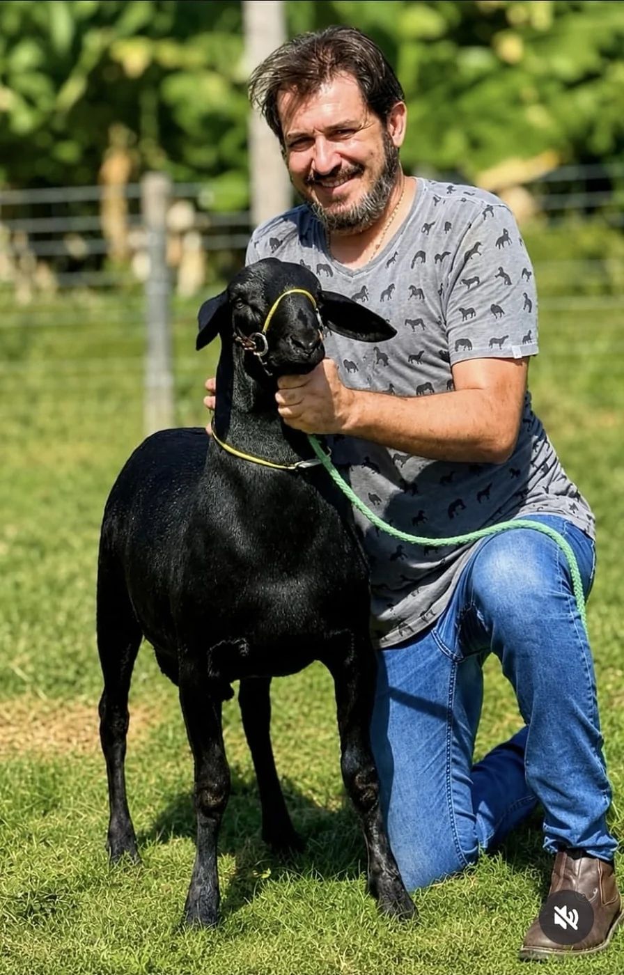 Augusto Cesinha ajoelhado no pasto, segurando pelo cabresto uma ovelha Santa Inês preta